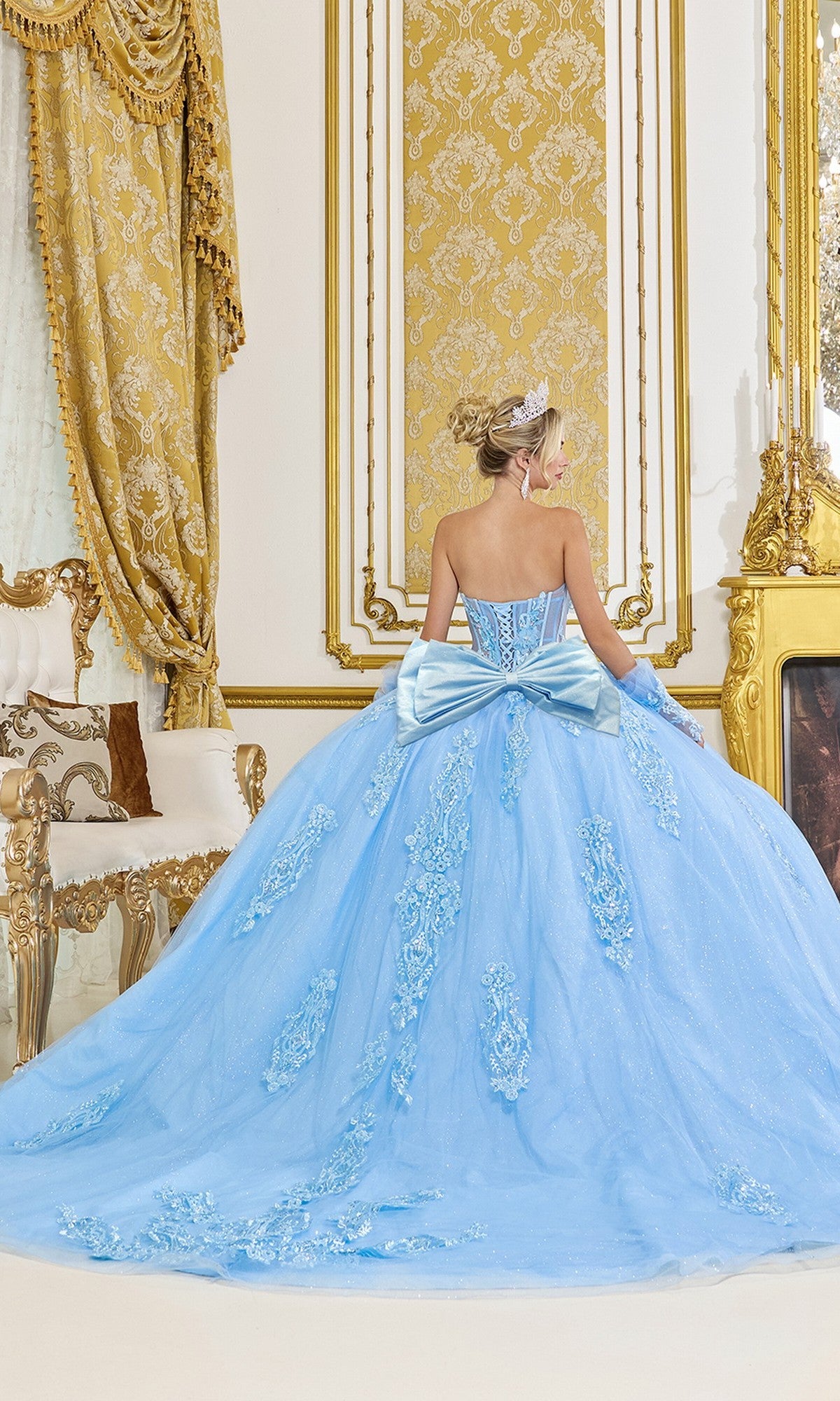 A woman in the Quinceanera Dress 5091 by Dancing Queen, a blue strapless gown with sequin appliquรฉ, stands facing away in an ornate, gold-accented room with decorative curtains and elegant furniture.